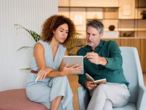 A woman and a man engaged in a discussion while reviewing a tablet, seated in a modern, stylish office space.