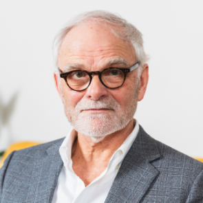 A man wearing glasses and sporting a grey beard sits on a bright yellow couch, creating a warm and inviting atmosphere.