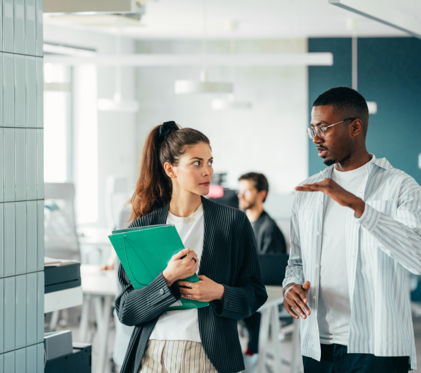 Two individuals engaged in a conversation within a professional office environment, surrounded by desks and office equipment.