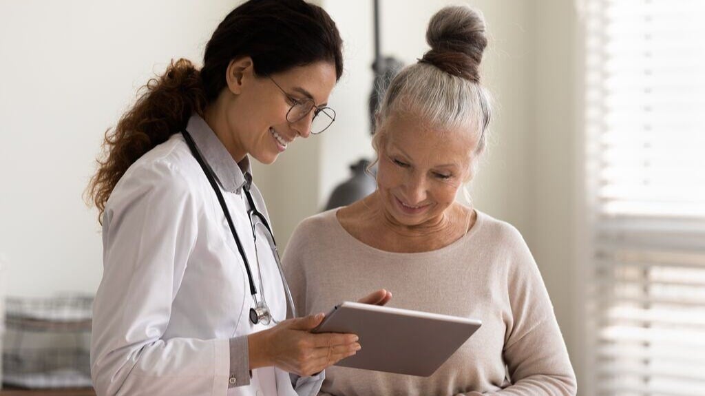 Happy-female-doctor-and-senior-patient-look-at-tablet